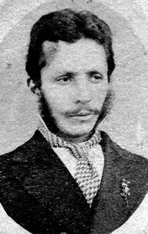 Старинна фотография на млад мъж с бакенбарди и мустаци, с тъмно сако и шарена връзка.
